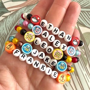 May include: A collection of five elastic bracelets featuring white letter beads spelling the names ETHAN, ALEX, LEO, JAKE, and FRANKIE. The bracelets include colourful round beads and flat circular charms depicting various cartoon characters and red pokeballs.
