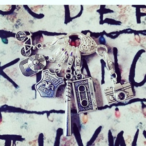 May include: Silver charm bracelet featuring a variety of charms, including a bicycle, a baseball bat, a police badge, a cassette tape, a camera, and a hat. The charms are attached to a silver chain with a heart-shaped clasp.