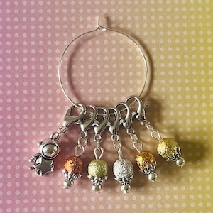 May include: A silver hoop with five silver stitch markers with beads. The beads are gold, copper, and silver with a glitter finish.