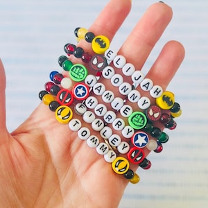 Könnte beinhalten: Ein Set aus acht bunten Perlenarmbändern mit Superhelden-Anhängern und weißen Buchstabenperlen, die die Namen Elijah, Sonny, Jamie, Harry, Finley und Tommy buchstabieren.