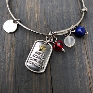 May include: A silver bangle bracelet with a silver charm that says "Duty Honor Loyalty Courage" with a gold star. The bracelet also has a red crystal, a white bead, and a blue stone charm.