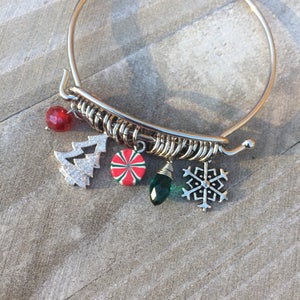 May include: A silver bangle bracelet with a variety of charms, including a silver snowflake, a green gem, a red and white candy cane, and a silver Christmas tree with red and white accents.