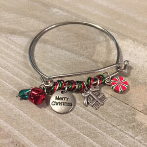 May include: A silver bangle bracelet with a red and green wire wrapped around it. The bracelet has several charms including a bell, a gift box, a candy cane, and a charm that says "Merry Christmas".