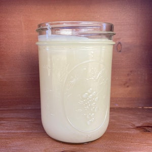 May include: A clear glass mason jar filled with a creamy, off-white substance. The jar has a metal lid and a decorative embossed design featuring leaves and grapes. The jar is sitting on a wooden surface.
