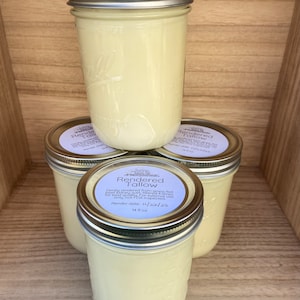 Puede incluir: Cuatro frascos de vidrio llenos de una sustancia cremosa de color amarillo p&aacute;lido, apilados sobre una estanter&iacute;a de madera. Los frascos tienen tapas plateadas y etiquetas que dicen "Rendered Tallow". Cada frasco contiene unos 414 ml.