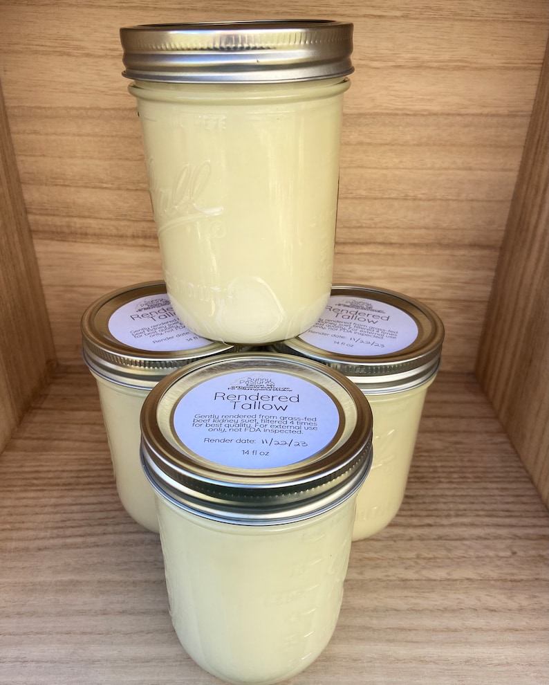 Puede incluir: Cuatro frascos de vidrio llenos de una sustancia cremosa de color amarillo p&aacute;lido, apilados sobre una estanter&iacute;a de madera. Los frascos tienen tapas plateadas y etiquetas que dicen "Rendered Tallow". Cada frasco contiene unos 414 ml.