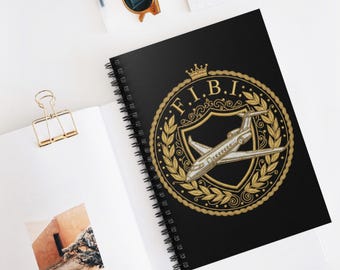 May include: Black spiral-bound notebook featuring a gold emblem with the letters "F.I.B.I." and a stylized airplane. The emblem is surrounded by a gold laurel wreath and a crown. The notebook is on a white surface.
