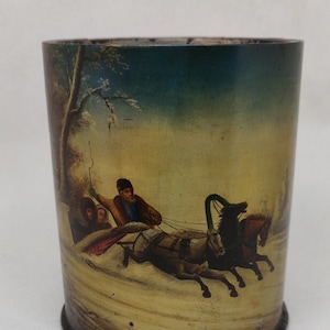 Op de afbeelding: Een vintage blikken doos met een geschilderd tafereel van een paardenslee in een besneeuwd winterlandschap. De slee wordt getrokken door twee paarden en er zitten drie mensen in. De blik is geschilderd in tinten blauw, groen, geel en bruin.