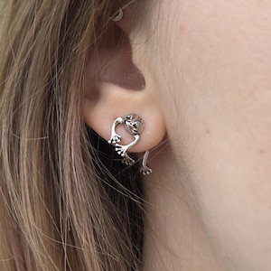 May include: Silver frog stud earrings. The earrings have a detailed frog design with outstretched legs. The earrings are worn on the ear of a person with light brown hair, against a blurred green backdrop.