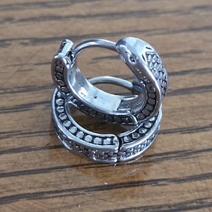 May include: A pair of silver hoop earrings. The earrings feature a detailed design with small, dark accents. The hoops are stacked, highlighting the intricate craftsmanship. The earrings are made of a shiny, metallic material, reflecting light.