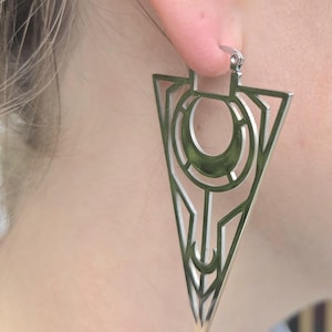 May include: A silver-colored geometric earring with a triangular shape. The earring features a cut-out design with a circular and crescent moon shape in the center. The earring is attached to a silver hoop.
