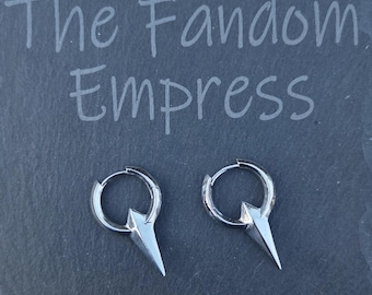 May include: A pair of silver hoop earrings with a pointed, dagger-like charm. The earrings are displayed on a dark gray surface, with the text "The Fandom Empress" etched above them.