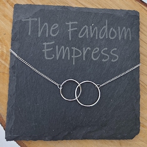 May include: A silver necklace featuring two interlocking circles, displayed on a dark gray slate. The text "The Fandom Empress" is etched into the slate. The necklace has a delicate chain.