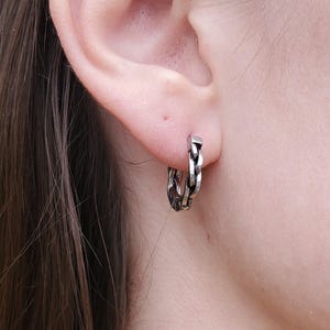 May include: A silver-coloured hoop earring with a chain-link design is worn on an ear. The earring is a small, circular shape with a textured surface. The earring is made of metal and has a shiny finish. The earring is a piece of jewellery.