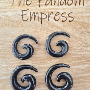 May include: Four black spiral-shaped ear gauges are displayed on a light brown wooden surface. The gauges have a glossy finish and a smooth, curved design. The text "The Fandom Empress" is written above the gauges.