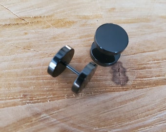 May include: Two black, circular stud earrings with a post and backing, displayed on a light brown wooden surface. The earrings have a simple, minimalist design, reflecting a modern aesthetic. The earrings are likely made of metal.