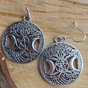 May include: A pair of silver earrings. Each earring has a circular pendant with a triple moon design, a pentagram, and Celtic knotwork. The earrings have a hook closure. The earrings are silver in colour.