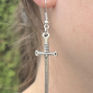May include: A silver-coloured sword-shaped earring with a hook closure. The earring features a detailed hilt and a long, pointed blade. The earring is worn on the ear, with the background out of focus.