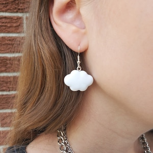 Peut inclure: Boucles d'oreilles pendantes en forme de nuage blanc avec des crochets argentés. Les boucles d'oreilles sont portées par une personne aux cheveux longs et bruns.
