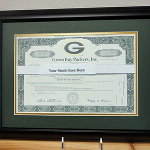Peut inclure: Un certificat d'actions encadré pour les Green Bay Packers, Inc. Le certificat a une bordure verte et dorée avec un logo 'G' vert et doré dans le coin supérieur gauche. Le texte 'Your Stock Goes Here' est imprimé sur une bannière blanche au milieu du certificat.