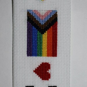 Puede incluir: Marcapáginas blanco de punto de cruz con una cinta arcoíris. El marcapáginas presenta una bandera del Orgullo bordada, un corazón rojo y las palabras "Love is Love" en negro.
