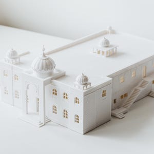 Puede incluir: Maqueta arquitectónica blanca de un edificio con múltiples cúpulas, puertas arqueadas y pequeñas ventanas. El modelo tiene una base rectangular y una escalera que conduce a un segundo nivel. Las ventanas parecen tener un brillo amarillo cálido.