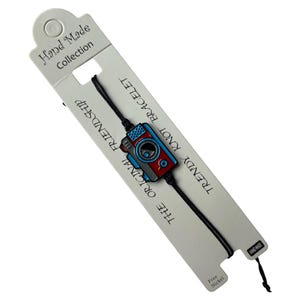 Peut inclure: Un bracelet d'amitié noir avec un charme de caméra rouge, bleu et blanc. Le bracelet est étiqueté "Trendy Knot" et "The Original Friendship Bracelet".