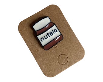 Könnte beinhalten: Eine kleine Emaille-Anstecknadel in Form eines Nutella-Glases mit braunem Körper, weißem Deckel und einem weißen Etikett, auf dem das Wort nutela in schwarzer Schrift gedruckt ist. Die Nadel ist auf einer braunen Kartonkarte befestigt.
