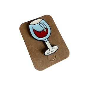May include: A metal enamel pin shaped like a wine glass with a light blue bowl, a white stem and base, and a deep red liquid inside. The pin is attached to a rectangular brown cardboard backing card with rounded corners and a small circular cutout.