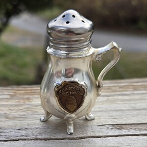 May include: Silver salt shaker with a brown Mammoth Cave National Park emblem. The shaker has a lid with holes and a handle.