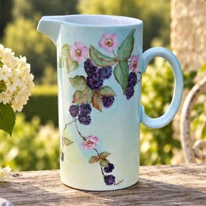 May include: A light blue ceramic pitcher featuring a hand-painted botanical design of dark purple blackberries, green leaves, and small pink flowers. The cylindrical vessel has a curved handle on the right side and a pouring spout on the top left.