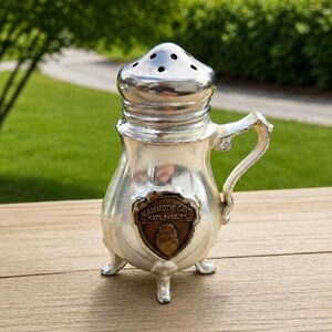 May include: A silver-toned metal shaker with a perforated dome lid, a curved handle, and three small feet. A bronze-colored shield emblem is attached to the front, featuring the embossed text MAMMOTH CAVE NATL PARX KY and a central graphic.