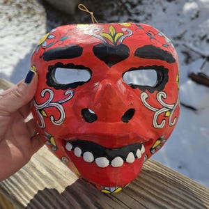 Op de afbeelding: Een rood papieren masker met zwarte en witte details. Het masker heeft een brede, tanden glimlach en bloemmotieven rond de ogen.