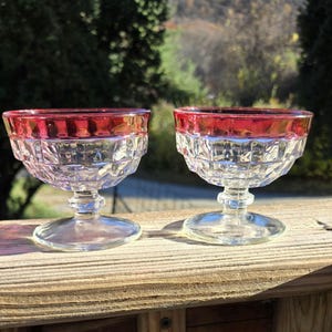 May include: Two clear glass dessert dishes with a red rim. The dishes have a diamond pattern and are on a wooden surface.