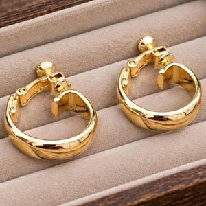 May include: A pair of polished gold-tone hoop earrings featuring a screw-back clip mechanism. The earrings have a smooth, rounded surface and a circular shape, designed for non-pierced ears, showcasing a reflective metallic finish throughout.