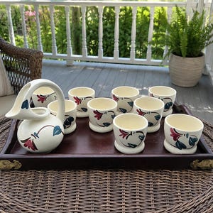 Könnte beinhalten: Ein Vintage-Set aus cremefarbenem Keramikkrug und passenden Tassen auf einem dunklen Holztablett. Der Krug hat einen gebogenen Griff und Ausguss. Die Tassen und der Krug sind mit roten, blauen und schwarzen floralen Designs verziert. Das Tablett hat Messing-Eckakzente.