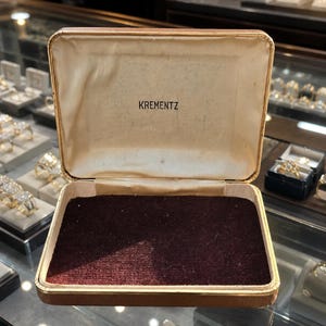 May include: A rectangular jewelry box with a brown exterior and a gold-toned metal trim. The interior lid is lined with off-white fabric featuring the brand name KREMENTZ printed in black. The base is lined with a dark burgundy velvet material.