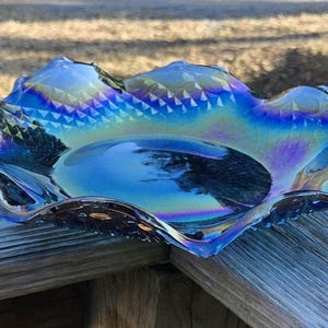May include: Iridescent blue glass bowl with a ruffled edge. The bowl has a central depression and a textured, geometric pattern around the rim. The glass reflects a rainbow of colors, including blue, purple, and green.