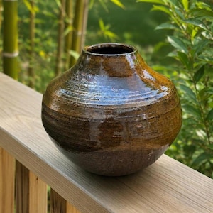 May include: A round, wide-bodied ceramic vase with a tapered neck and a small circular opening. The surface features a glossy, mottled brown glaze with horizontal ridges and subtle variations in color, ranging from dark espresso to golden amber.