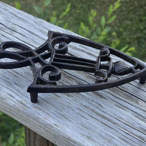 May include: A black cast iron trivet with an ornate, scrolled design. The trivet has a flat base with three small feet and is designed to protect surfaces from hot cookware. The decorative piece is made of metal and is suitable for kitchen use.