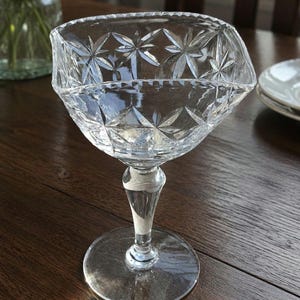 May include: A clear, cut-glass goblet with a starburst pattern on the bowl. The goblet has a faceted stem and a round base. The glass is sitting on a dark wooden table. The goblet is likely used for serving beverages.