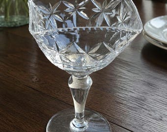 Peut inclure: Un gobelet en verre taillé transparent avec un motif d'éclat d'étoile sur le bol. Le gobelet a une tige facettée et une base ronde. Le verre est posé sur une table en bois sombre. Le gobelet est probablement utilisé pour servir des boissons.