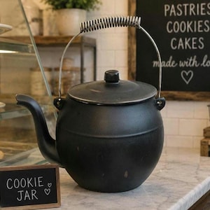 Pode incluir: Um pote de biscoitos preto e redondo com tampa e alça de mola. O pote tem um bico e está sobre uma bancada de mármore. Uma pequena placa de ardósia diz "COOKIE JAR".
