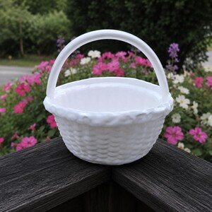 May include: A white ceramic basket with a woven texture and a curved handle. The basket is sitting on a dark wooden surface, with a background of pink and white flowers and green foliage.