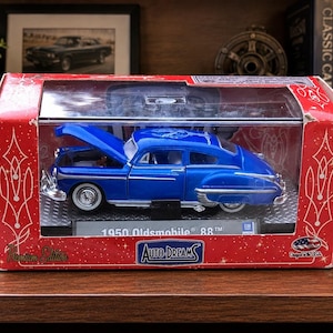 May include: A blue 1950 Oldsmobile 88 model car with its hood open, displayed in a red box. The box has a clear plastic front and features the text "1950 Oldsmobile 88" and "Auto-Dreams". The car has white-walled tires.