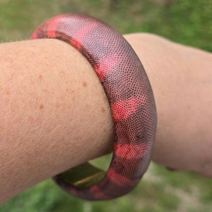 May include: A thick, round bangle bracelet with a textured surface. The bracelet features a pattern of red and brown hues. The accessory is worn on the arm, showcasing its design and color variations. The bracelet's design is reminiscent of reptile skin.