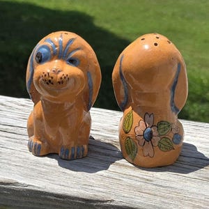May include: A pair of ceramic dog-shaped salt and pepper shakers. The shakers are orange with blue accents and feature a winking dog design on the front. The back of one shaker has a floral design.