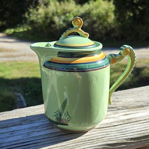 May include: A light green ceramic pitcher with a lid. The pitcher has a curved handle and spout. The lid is decorated with yellow and blue stripes. The pitcher is decorated with a painted plant design. The pitcher is sitting on a wooden surface.