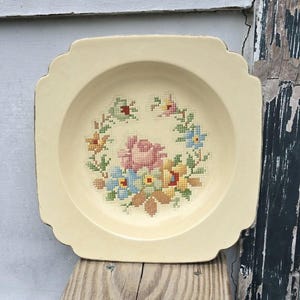 May include: A square, cream-colored ceramic plate with a raised rim and a floral design in the center. The design features a pink rose surrounded by yellow, blue, and green flowers. The plate has a gold trim around the edge.