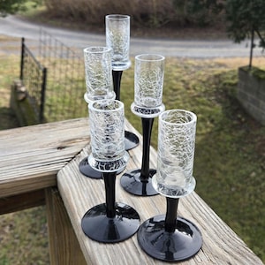 May include: Five clear glass shot glasses with black bases. The glasses have a crackled texture and are arranged in a row on a wooden surface.